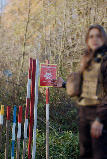 фонове зображення: жінка сапер вказує на знак небезпеки та дерев'яні кілки для маркування місцевості